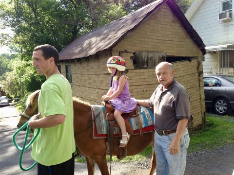 A young girl wearing a colorful helmet sits on a brown pony being led by two men in front of a rustic wooden shed.