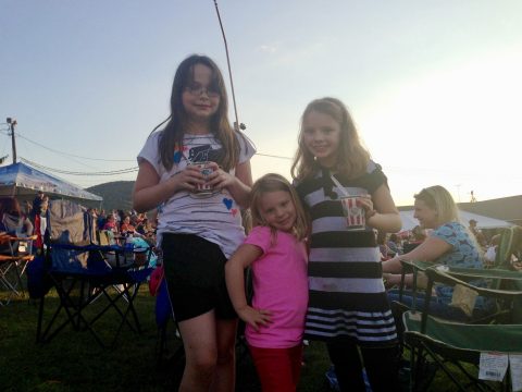 Three young girls standing outdoors at a festival or event, holding drinks in paper cups.