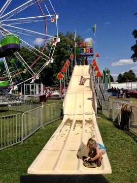 Two children on a long outdoor slide at a fair with a Ferris wheel in the background.
