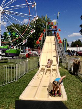 Two children playing on a large outdoor slide at a fair with a Ferris wheel in the background.