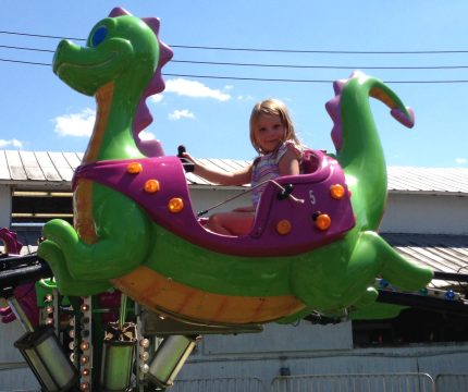 A young girl with blonde hair smiles while riding a colorful dragon-shaped amusement park ride on a sunny day.