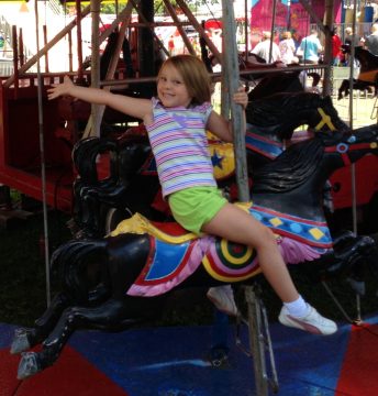 A smiling young girl with light brown hair wearing a striped shirt and green shorts rides a black carousel horse at an outdoor fair.
