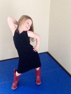 A young girl with blonde hair wearing a black dress and red boots posing on a blue mat indoors.
