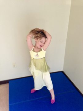 A young girl in a yellow and green dress stands on a blue mat with her hands behind her head.
