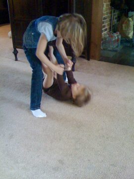 Two children playing on a carpeted floor; one child stands while the other lies on their back.