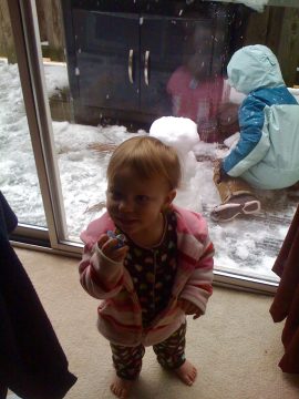A toddler in a pink striped jacket stands indoors holding a small object, looking toward the camera while snow falls outside through a sliding glass door.