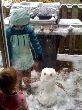 Two children in winter coats look at a small snowman with orange eyes in a snowy backyard.