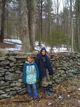 Two children in winter jackets standing in front of a stone wall in a wooded area with snow on the ground.