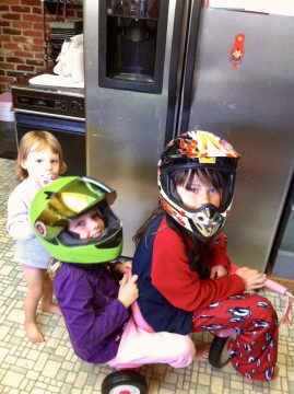 Three young children wearing colorful helmets pose in a kitchen with stainless steel appliances.