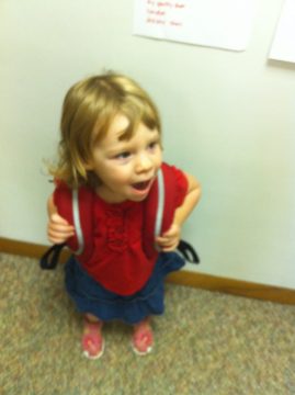 A young child with blonde hair wearing a red top and blue skirt stands indoors holding the straps of a small backpack.