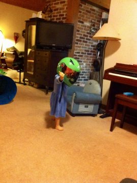 A small child in a blue dress wearing a large green motorcycle helmet stands indoors pointing forward.