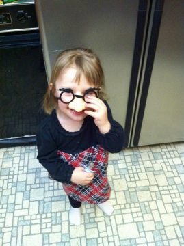 A young child wearing oversized glasses and a plaid dress stands in a kitchen holding a small object.
