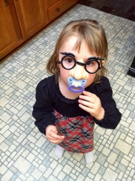 A young child wearing oversized glasses and a pacifier stands on a tiled floor in front of wooden cabinets.
