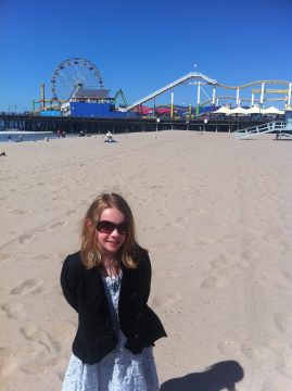 A young girl wearing sunglasses and a black jacket stands on a sandy beach with the Santa Monica Pier in the background.