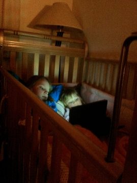 Two young children lying in a wooden crib at night, illuminated by a lamp above and looking toward a laptop screen.