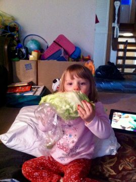 A young child in pink pajamas sits on a bed holding a large head of lettuce wrapped in plastic near their face.