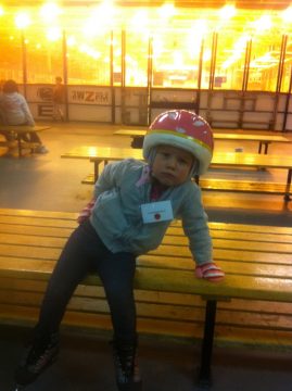 A young child wearing a pink helmet and roller skates sits on a wooden bench in an indoor skating rink.