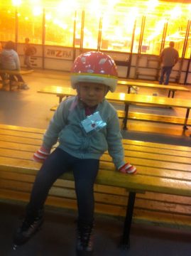 A young child wearing a red helmet and roller skates sits on a yellow bench in an indoor rink.