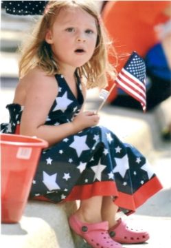 A young girl in a patriotic dress holding a small American flag and sitting next to a red bucket.