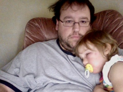 A man with glasses and a beard lies in a chair holding a sleeping child with a pacifier.