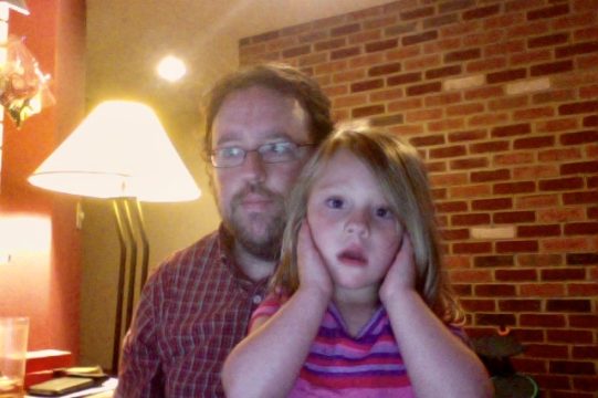 A man with glasses and a beard stands behind a young girl in a striped shirt who is holding her cheeks, both looking toward the camera indoors near a brick wall.