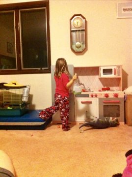 A young girl in pajamas stands at a toy kitchen holding a bottle while a stuffed animal lies on the floor nearby.