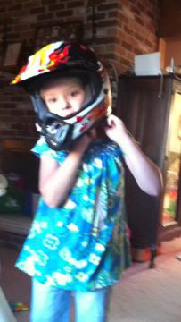 A young child wearing a colorful motocross helmet and blue patterned shirt stands indoors near a brick wall.
