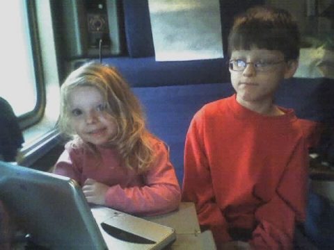 Two young children sitting on a train with a laptop in front of them.