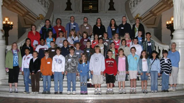 A group of children and adults pose for a photo on the steps of a grand building. A large group of children and adults posing for a photo on the steps of a grand building with white columns and statues.