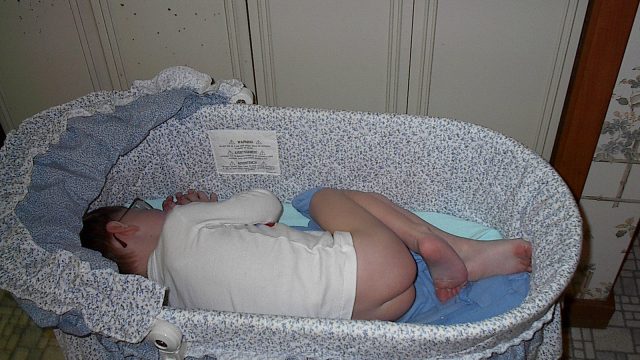 A person sleeping in a baby bassinet with blue and white patterned fabric.