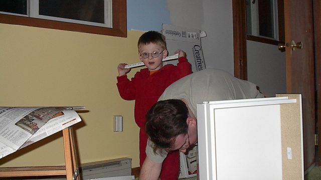 A young boy in red pajamas holds a ruler like a check while an adult works on furniture nearby.