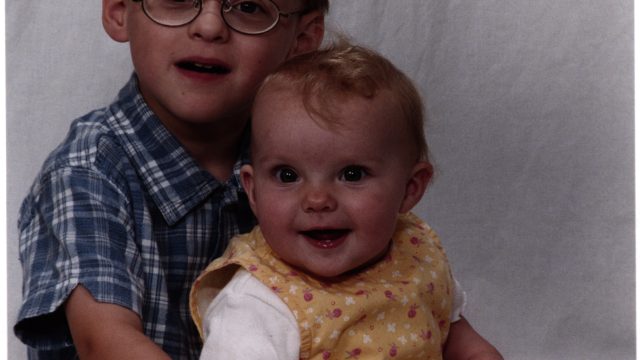 A young boy with glasses holding a smiling baby in a yellow dress against a white backdrop.