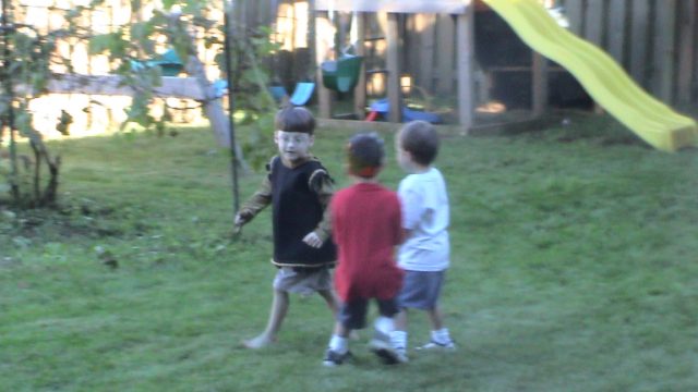 Three young boys playing in a grassy backyard with a wooden playset and yellow slide.