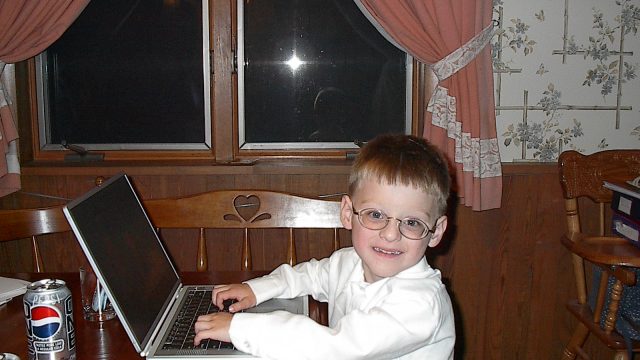 A young boy wearing glasses and a white shirt sits at a wooden table using a laptop with a Pepsi can nearby.