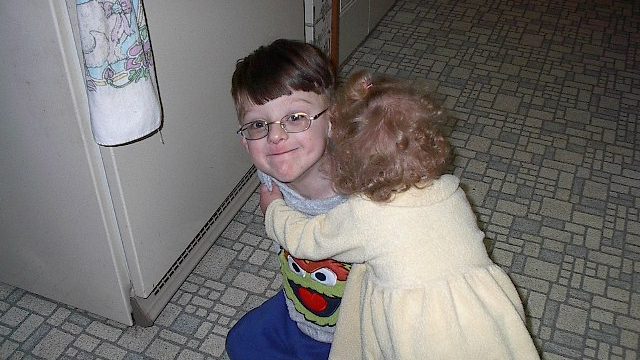 A young boy with glasses smiles while hugging a toddler in a yellow dress near a refrigerator.