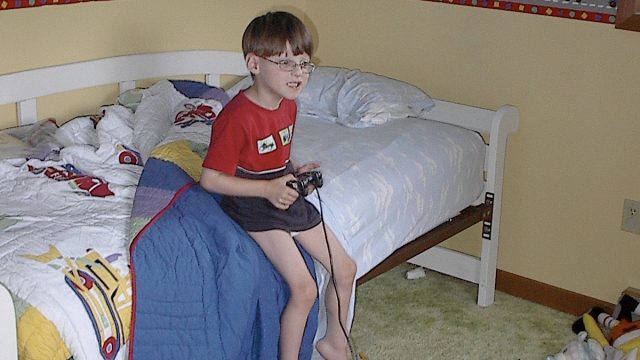 A young boy with glasses sits on a bed holding a video game controller in a messy bedroom.