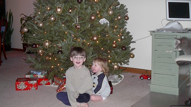 Two young children sitting on the floor in front of a decorated Christmas tree with wrapped gifts nearby.