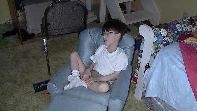 A young boy wearing glasses and a white t-shirt sits cross-legged in a light blue recliner chair with footrest, looking to the side.