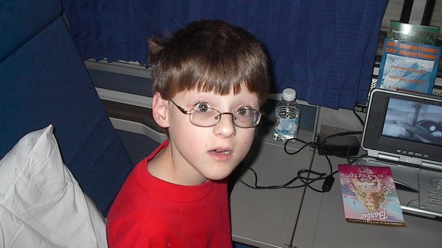 A young boy in a red shirt and glasses looks at the camera while seated on a train. A young boy with glasses wearing a red shirt sits on a train seat next to a small TV and water bottle.