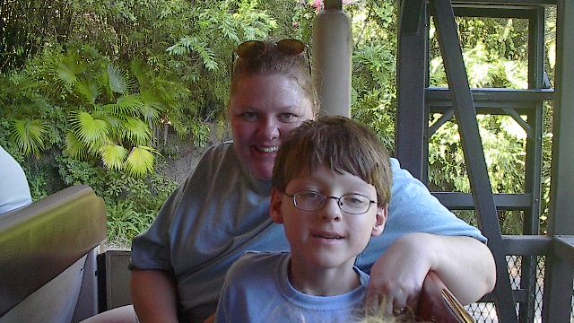 A smiling woman and a young boy enjoy a ride outdoors. A woman and a young boy with glasses sit together on a ride vehicle in a park setting.