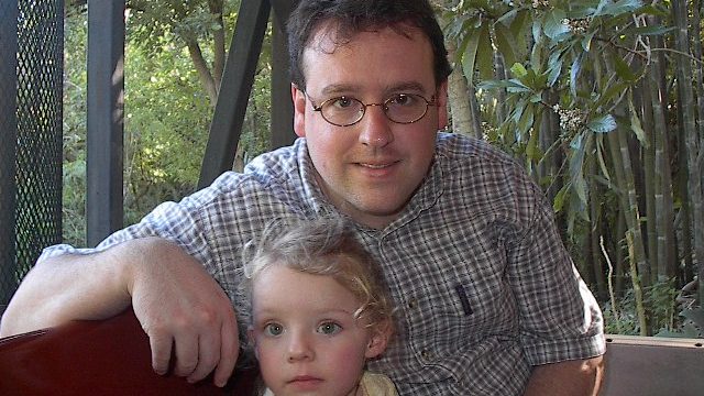 Man and child posing together outdoors. A man wearing glasses and a plaid shirt sits behind a young child with light hair in an outdoor setting.