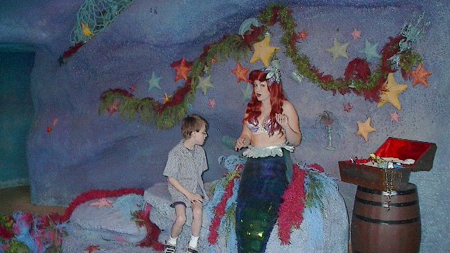 A child stands beside a mermaid figure in a festive, cave-like setting. A mermaid statue with red hair sits next to a child in a decorated underwater-themed room.