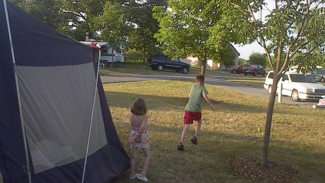 Children playing outside near a tent in a suburban setting. Two children standing on grass near a tent in a residential area with parked cars and trees.