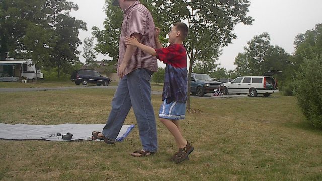 A man and a young boy walking outdoors on a grassy area with vehicles and trees in the background. A man in a striped shirt and jeans walks on grass while a young boy in a tie-dye shirt holds his arm.