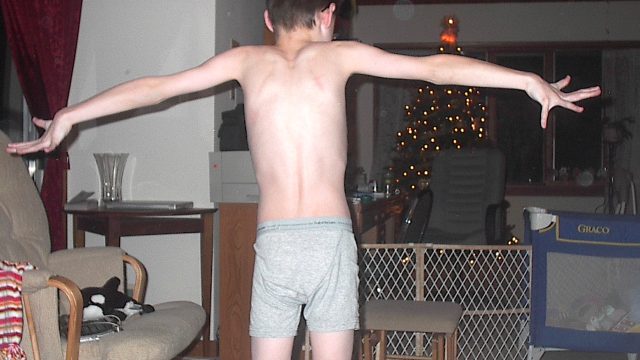 A shirtless boy in underwear stands with arms outstretched in a living room with Christmas lights and furniture.