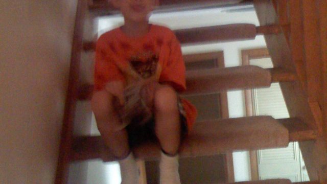 A young boy wearing glasses and an orange shirt sits on a carpeted wooden staircase in a home.