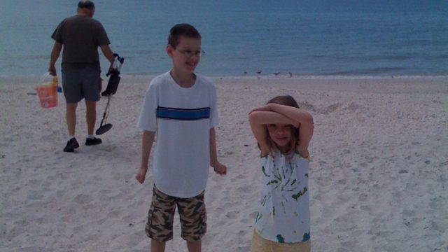 Children posing on a sunny beach near the ocean. Two children standing on a sandy beach with the ocean in the background; one child holds hands over eyes.
