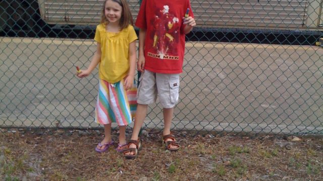 Two children pose in front of a train car near a chain-link fence. Two children standing in front of a train car with a chain-link fence behind them.