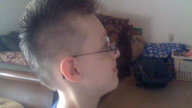 A young boy with spiky hair and glasses stands in profile looking upward inside a living room.