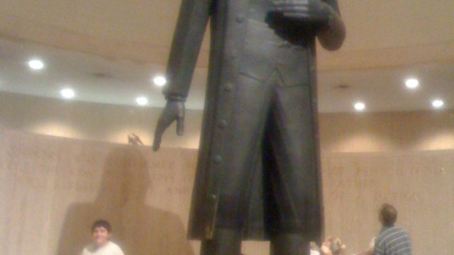 People gather near the base of a tall bronze statue in a domed interior space. A large bronze statue of a man in formal attire stands inside a circular room with people around its base.